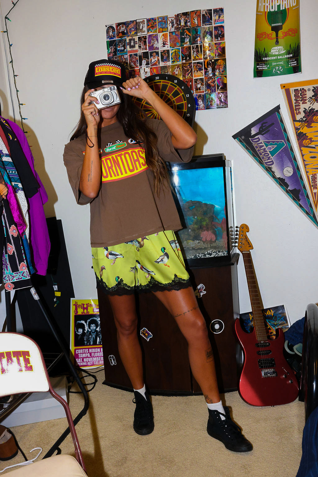 Model wearing brown shirt and green satin shorts, taking a picture in a room with posters and a guitar, frontal view