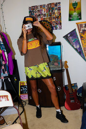 Model wearing brown shirt and green satin shorts, taking a picture in a room with posters and a guitar, frontal view
