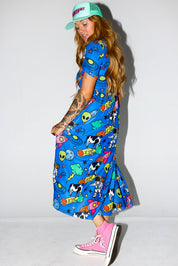 Model wearing a colorful blue dress with cartoon patterns, pink sneakers, and a green cap on a white background, side view