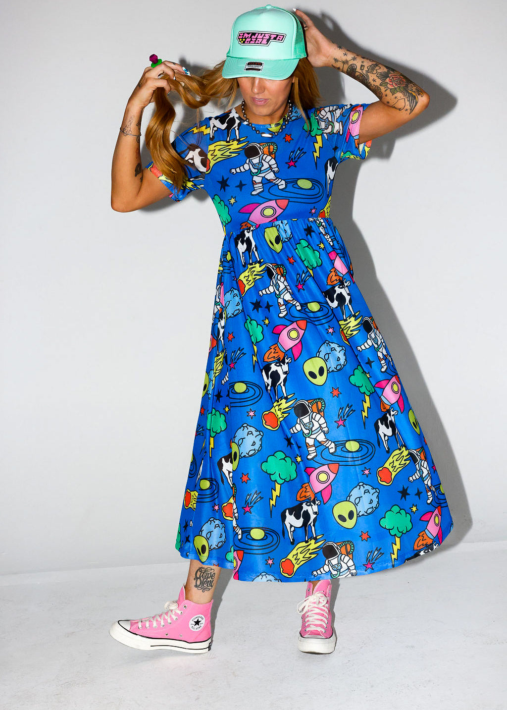 Model wearing a colorful blue dress with cartoon patterns, pink sneakers, and a green cap on a white background, frontal view