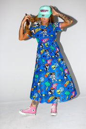 Model wearing a colorful blue dress with cartoon patterns, pink sneakers, and a green cap on a white background, frontal view