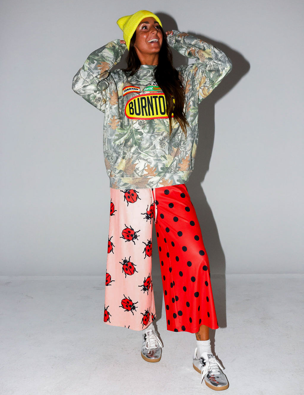 Model wearing a camo crewneck with Burnt Toast logo, red polka dot pants, and a yellow beanie, on a white background, frontal view