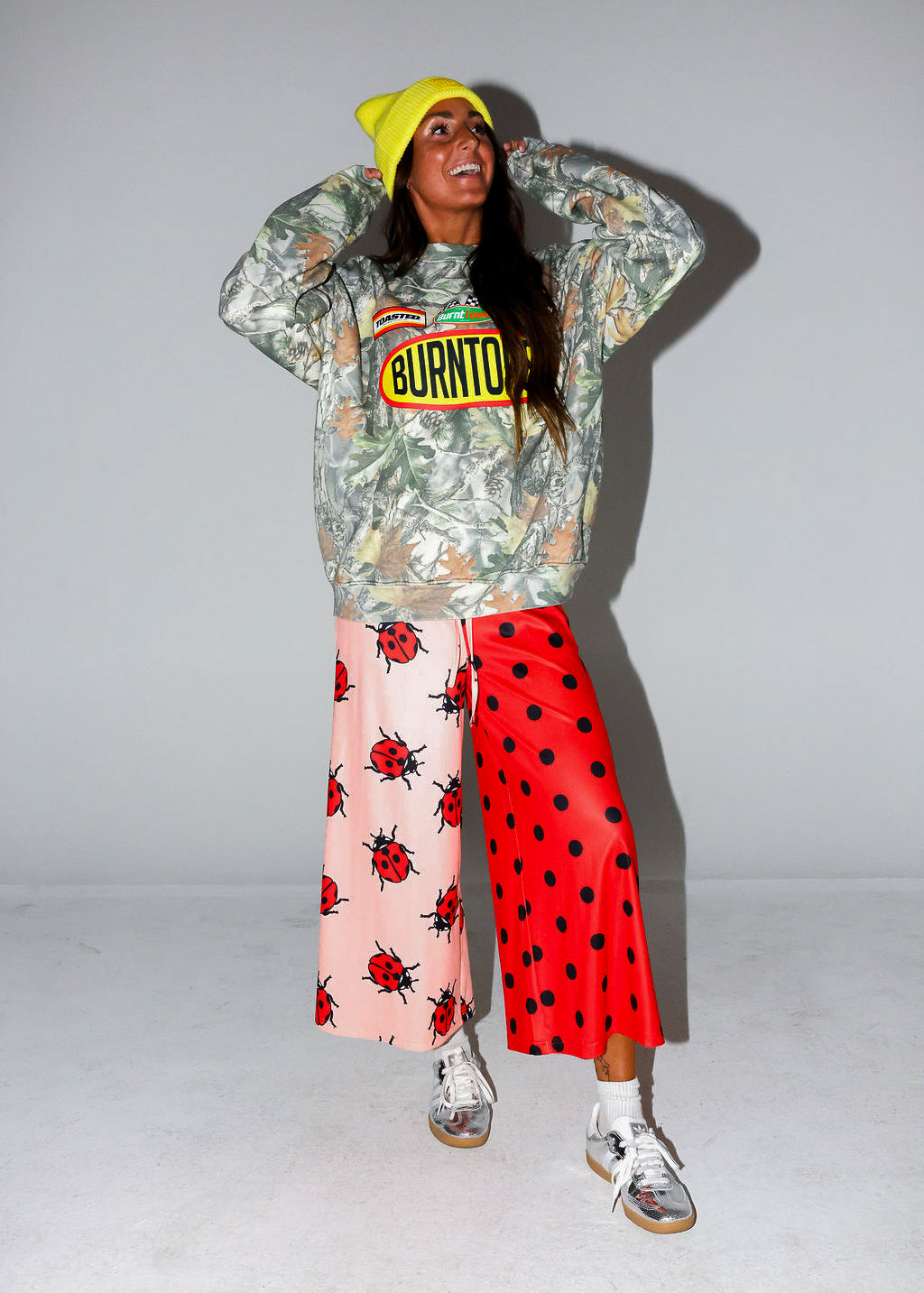 Model wearing a camo crewneck with Burnt Toast logo, red polka dot pants, and a yellow beanie, on a white background, frontal view