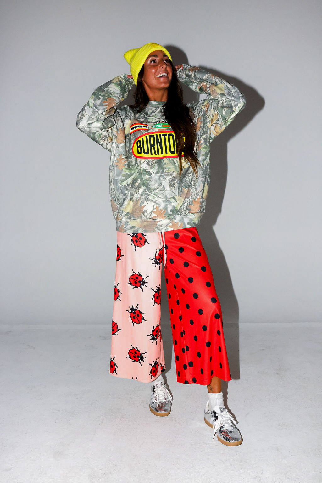 Model wearing a camo crewneck with Burnt Toast logo, red polka dot pants, and a yellow beanie, on a white background, frontal view