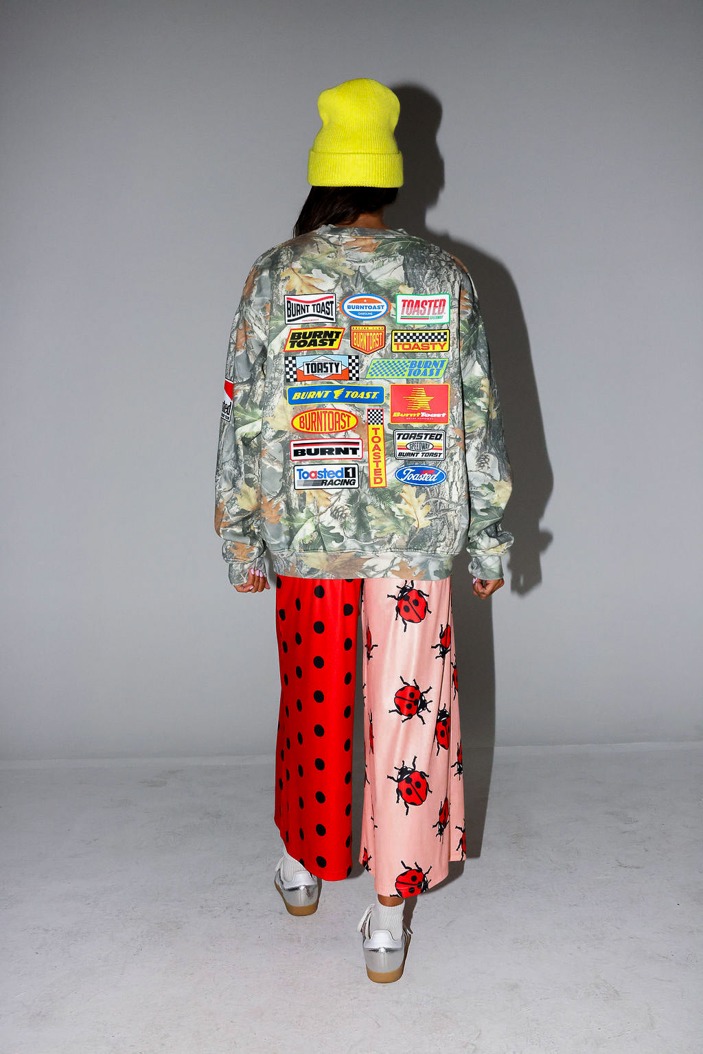 Model wearing a camo crewneck with logos, red polka dot pants, and a yellow beanie, on a white background, back view