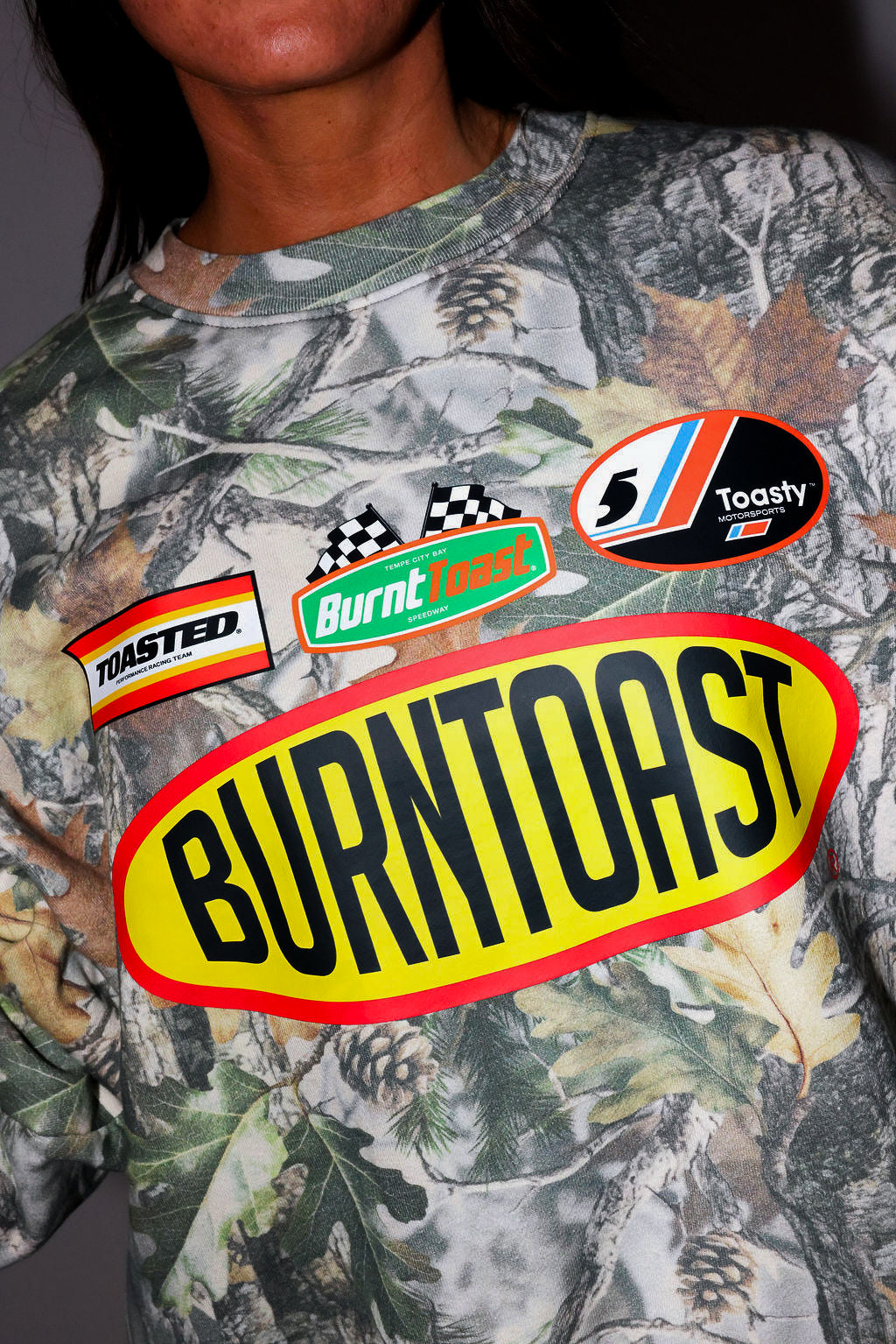 Model wearing a camouflage crewneck with 'Burntoast' branding, and other logos, frontal view