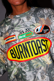 Model wearing a camouflage crewneck with 'Burntoast' branding, and other logos, frontal view
