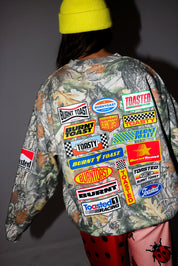 Model wearing a neon yellow beanie, camouflage crewneck with various brand logos on a dark background, back view