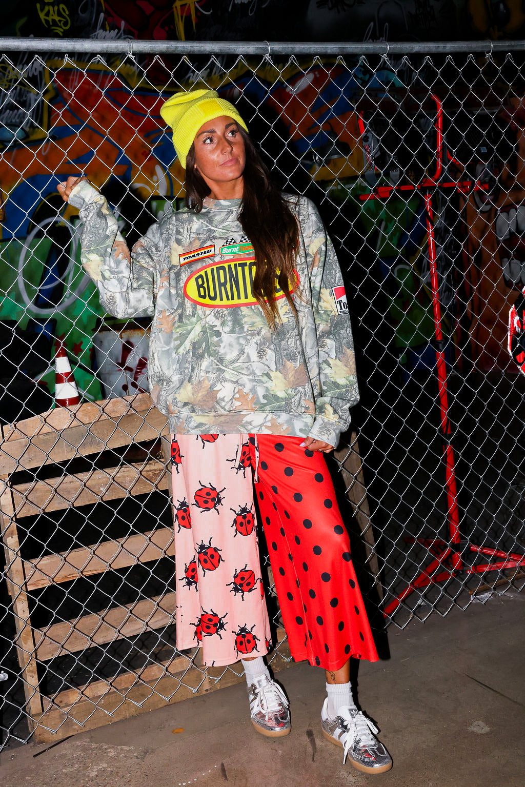 Model wearing a camo crewneck with Burnt Toast logo, red polka dot pants, and a yellow beanie, standing in front of a chain link fence, frontal view