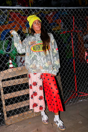 Model wearing a camo crewneck with Burnt Toast logo, red polka dot pants, and a yellow beanie, standing in front of a chain link fence, frontal view