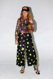 Model wearing a colorful patterned shirt and black pants with yellow stars on a white background, frontal view
