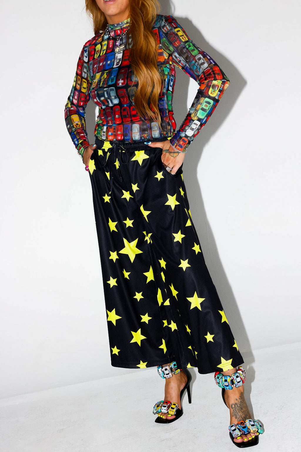 Model wearing a colorful patterned shirt and black pants with yellow stars on a white background, frontal view