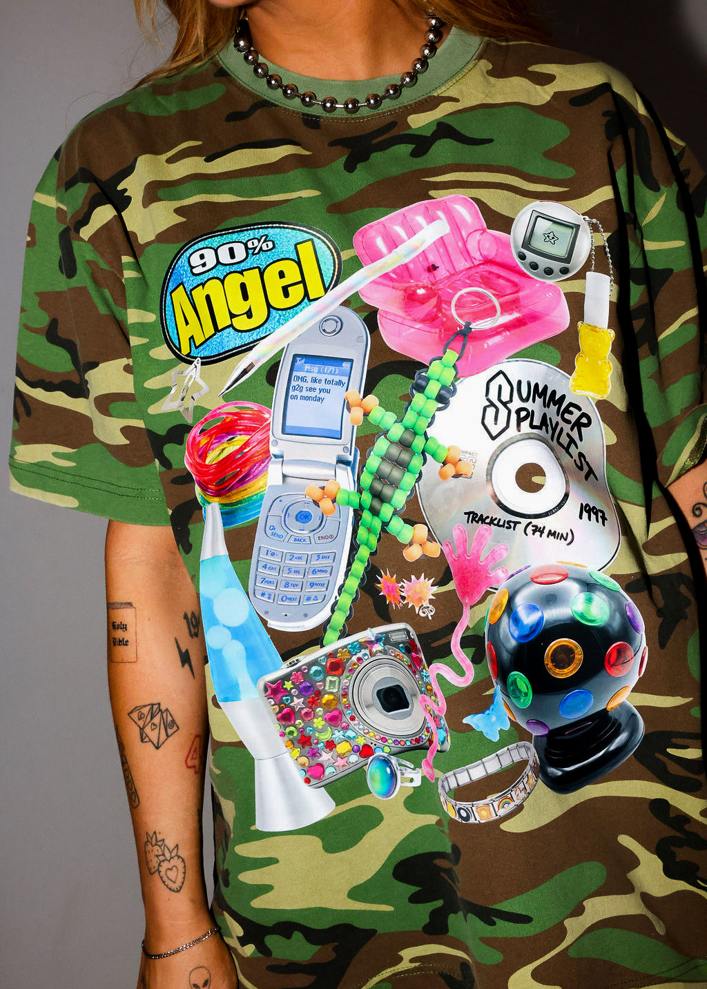 Model wearing camouflage t-shirt with colorful graphic designs and text on a plain background, close up frontal view