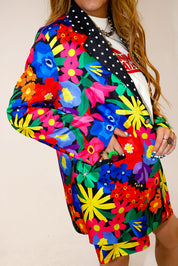 Model wearing a colorful floral jacket with polka dot collar, and matching shorts, with hand in pocket, a plain background, side view