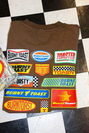 Brown t-shirt with multiple colorful brand logos on a checkered floor.