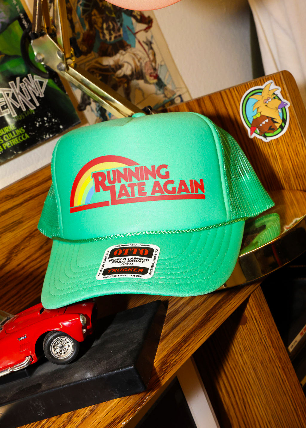 Green trucker with 'Running Late Again' text on a wooden surface with a red car model and other items, frontal view