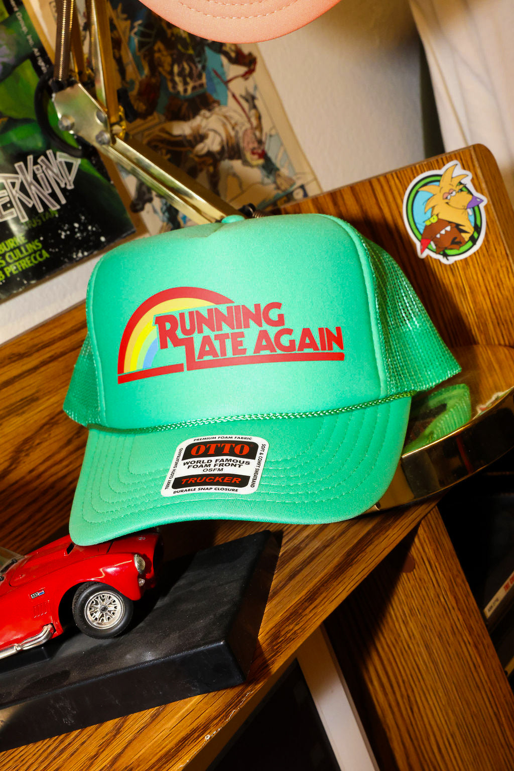 Green trucker with 'Running Late Again' text on a wooden surface with a red car model and other items, frontal view