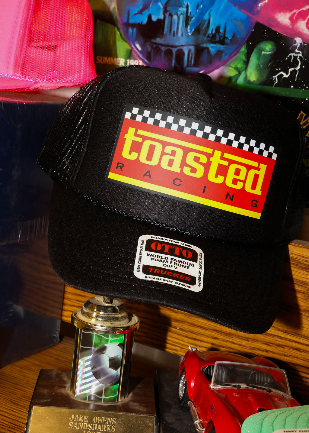 Black trucker with 'Toasted Racing' logo on a wooden surface with colorful items in the background, frontal view