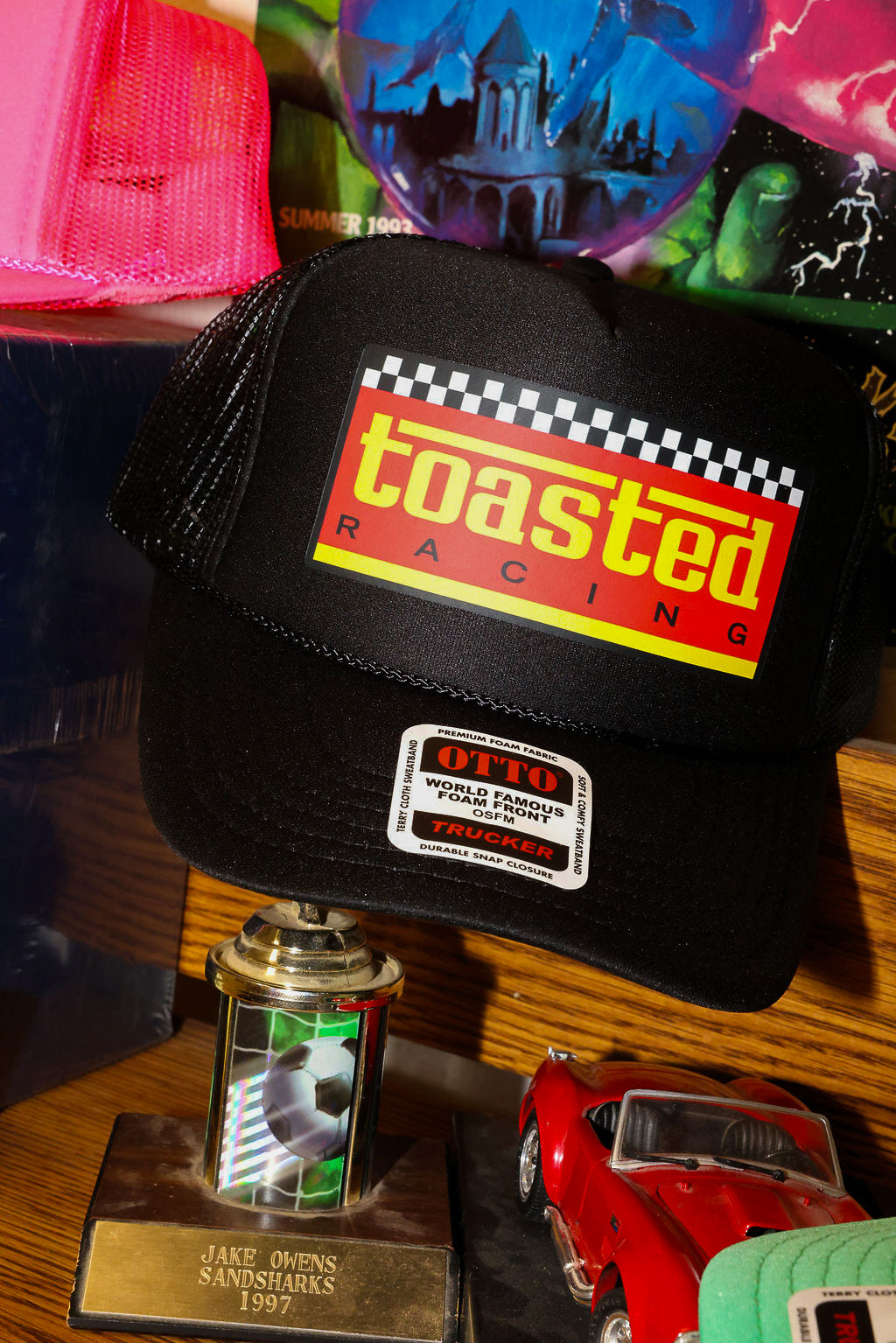 Black trucker with 'Toasted Racing' logo on a wooden surface with colorful items in the background, frontal view