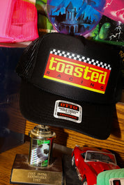 Black trucker with 'Toasted Racing' logo on a wooden surface with colorful items in the background, frontal view