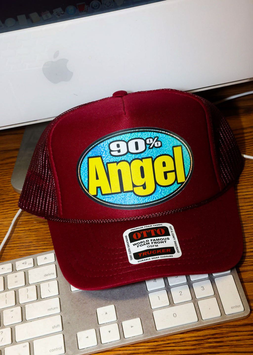Maroon trucket hat with '90% Angel' text on a desk with a computer monitor and keyboard, frontal view