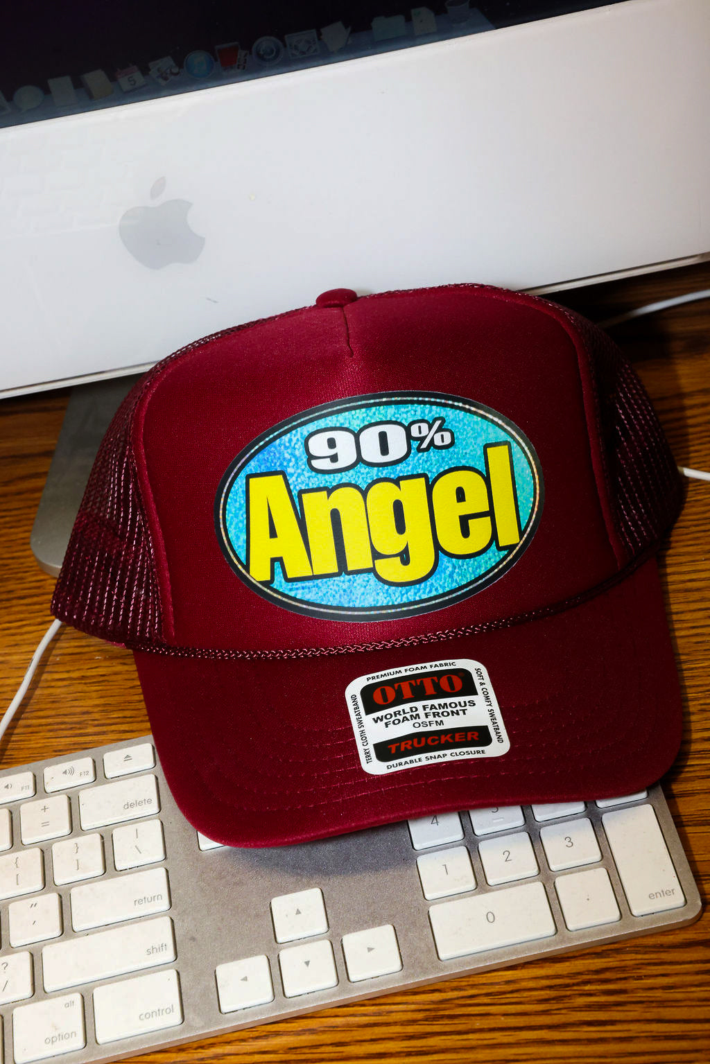 Maroon trucket hat with '90% Angel' text on a desk with a computer monitor and keyboard, frontal view