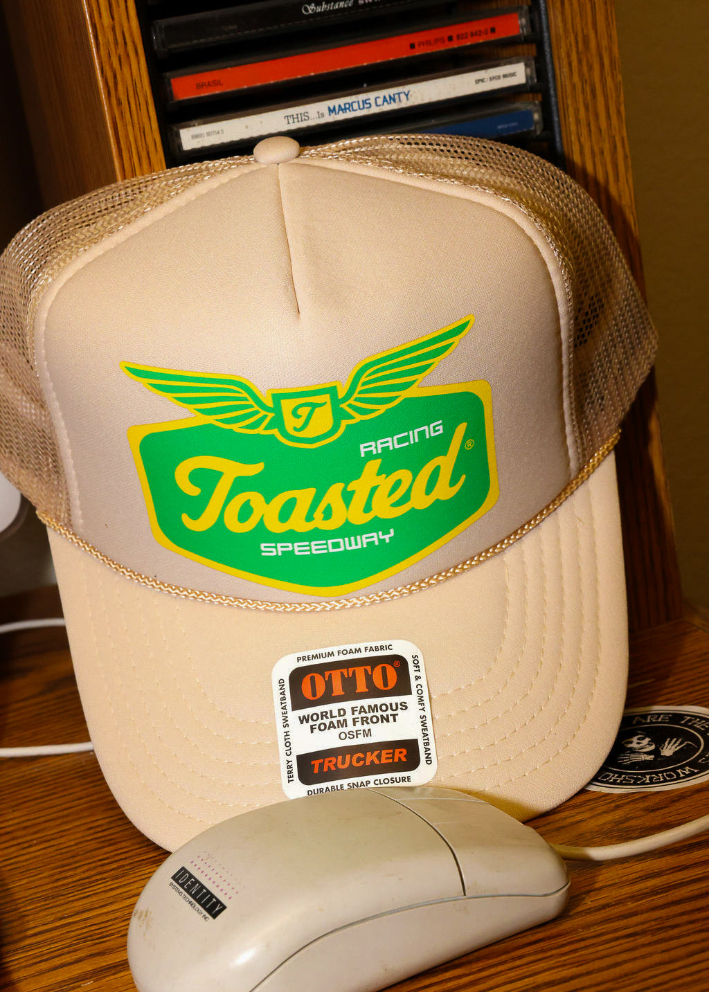 Tan trucker hat with 'Toasted Speedway' logo and a computer mouse on a wooden surface. frontal view