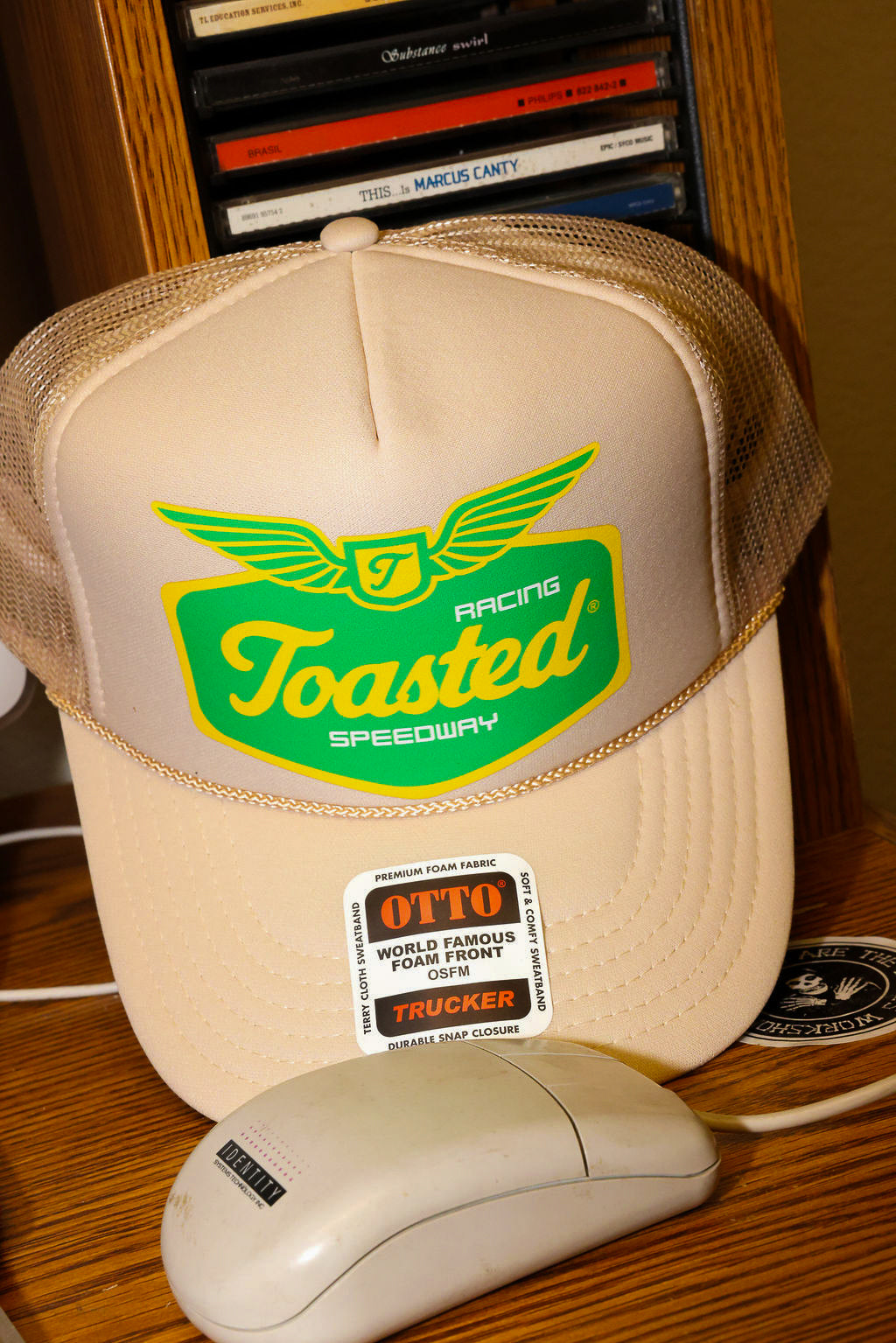 Tan trucker hat with 'Toasted Speedway' logo and a computer mouse on a wooden surface. frontal view