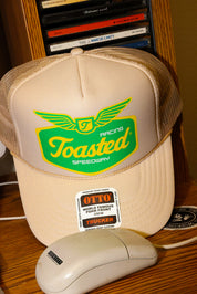Tan trucker hat with 'Toasted Speedway' logo and a computer mouse on a wooden surface. frontal view