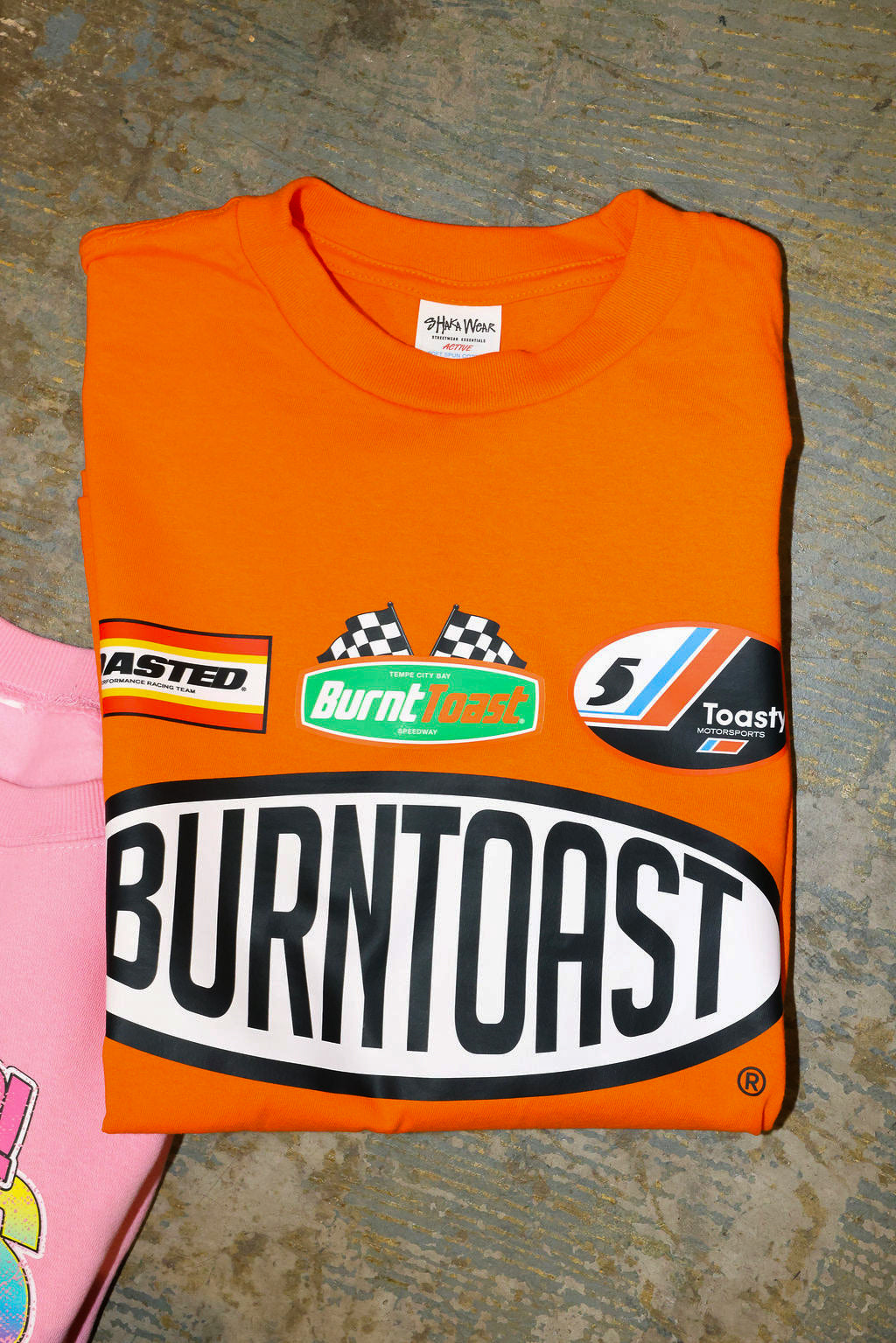 Orange t-shirt with 'Burntoast' branding on a textured surface, flat lay frontal view
