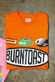 Orange t-shirt with 'Burntoast' branding on a textured surface, flat lay frontal view