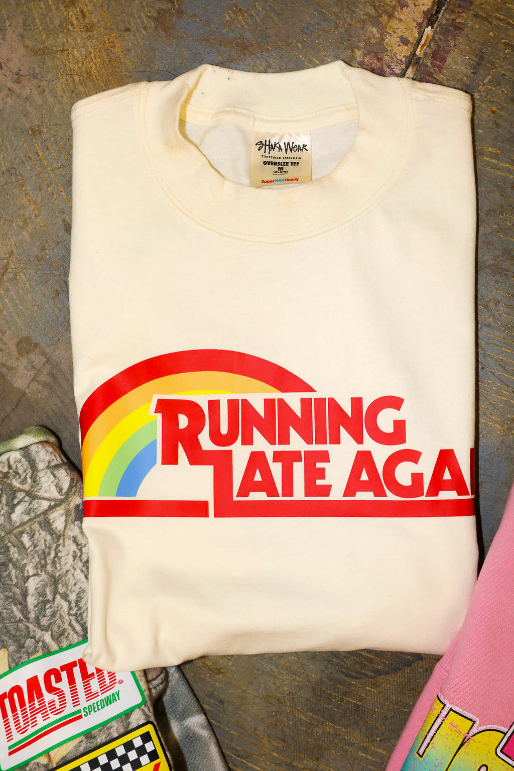 Folded off-white shirt with 'Running Late Aga' graphic on a textured surface, flay lay frontal view
