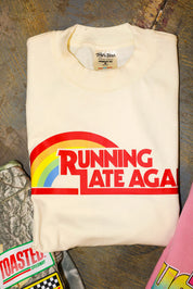Folded off-white shirt with 'Running Late Aga' graphic on a textured surface, flay lay frontal view