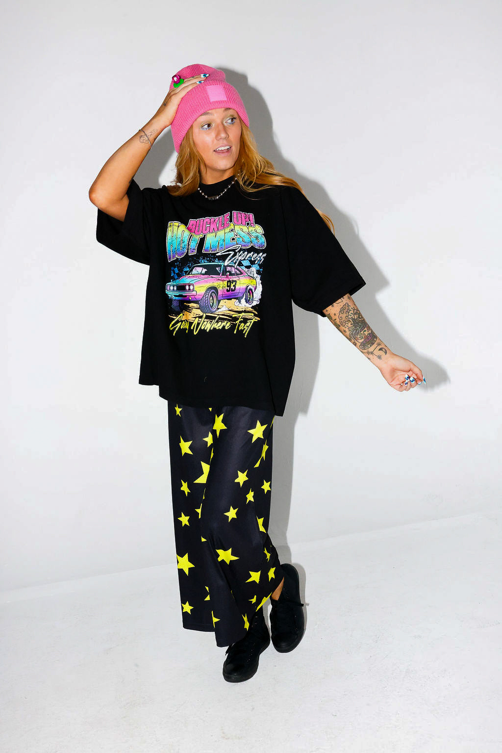 Model wearing a black short sleeve shirt with colorful graphic design and a pink beanie, and black and yellow pants, on a white background, frontal view