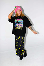 Model wearing a black short sleeve shirt with colorful graphic design and a pink beanie, and black and yellow pants, on a white background, frontal view