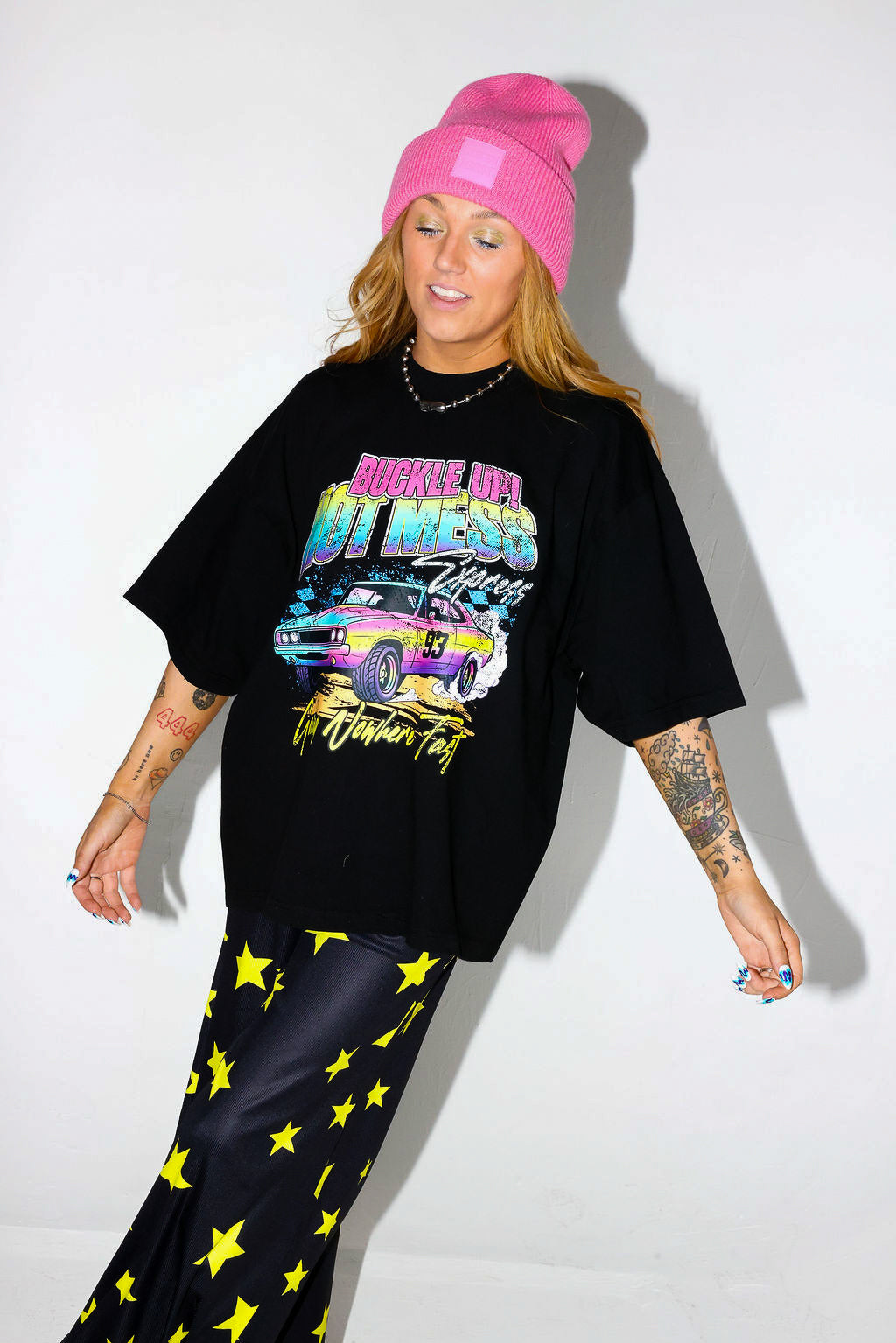 Model wearing a black short sleeve shirt with colorful graphic design and a pink beanie, and black and yellow pants, on a white background, close up frontal view