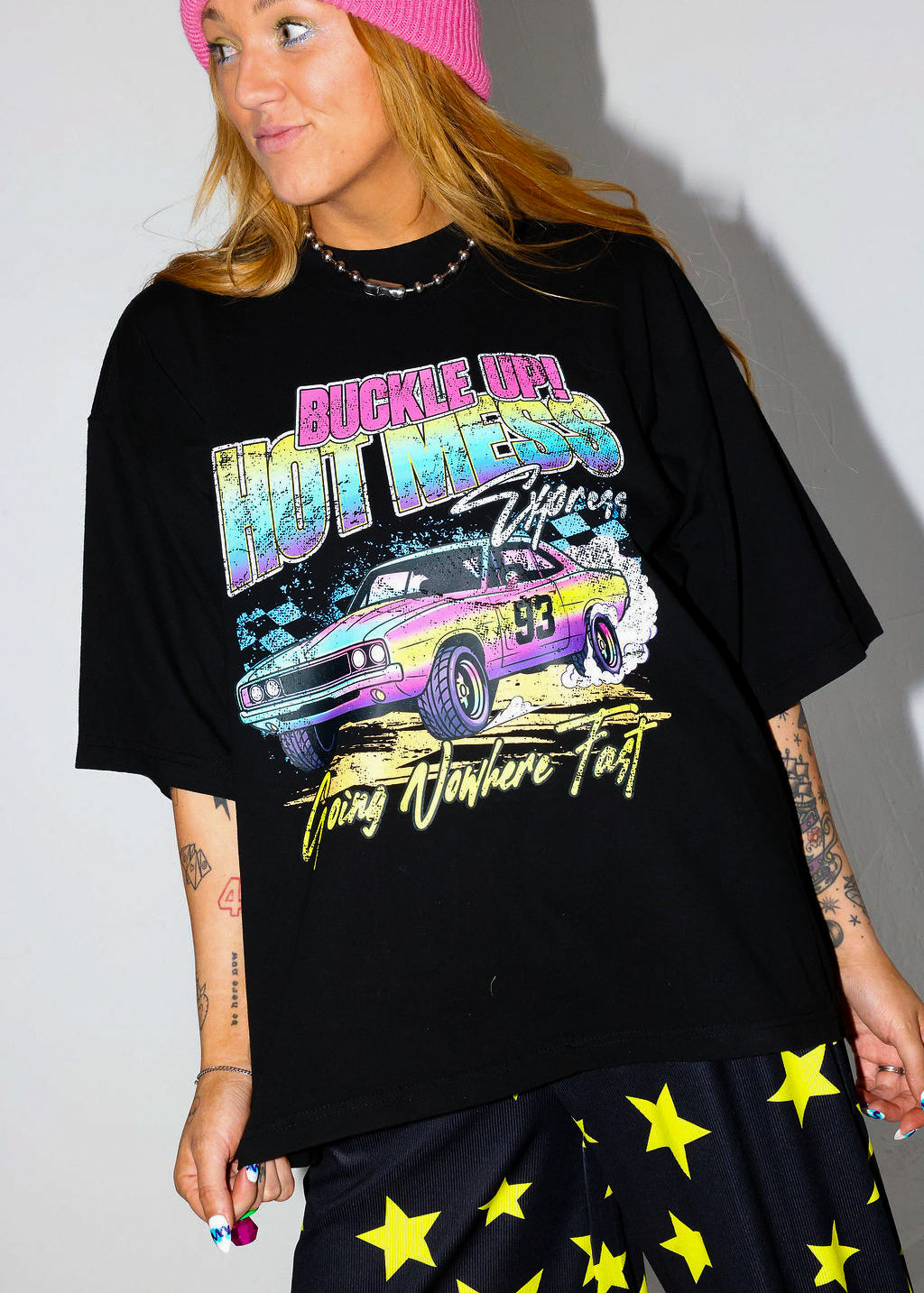 Model wearing a black short sleeve shirt with colorful graphic design and a pink beanie, and black and yellow pants, on a white background, close up frontal view