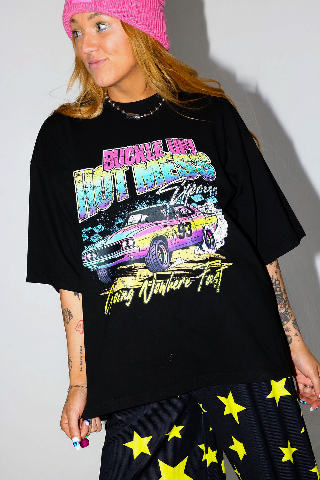Model wearing a black short sleeve shirt with colorful graphic design and a pink beanie, and black and yellow pants, on a white background, close up frontal view