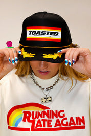Model wearing black TOASTED trucker hat, an ivory shirt with colorful text, against a plain background, close up frontal view