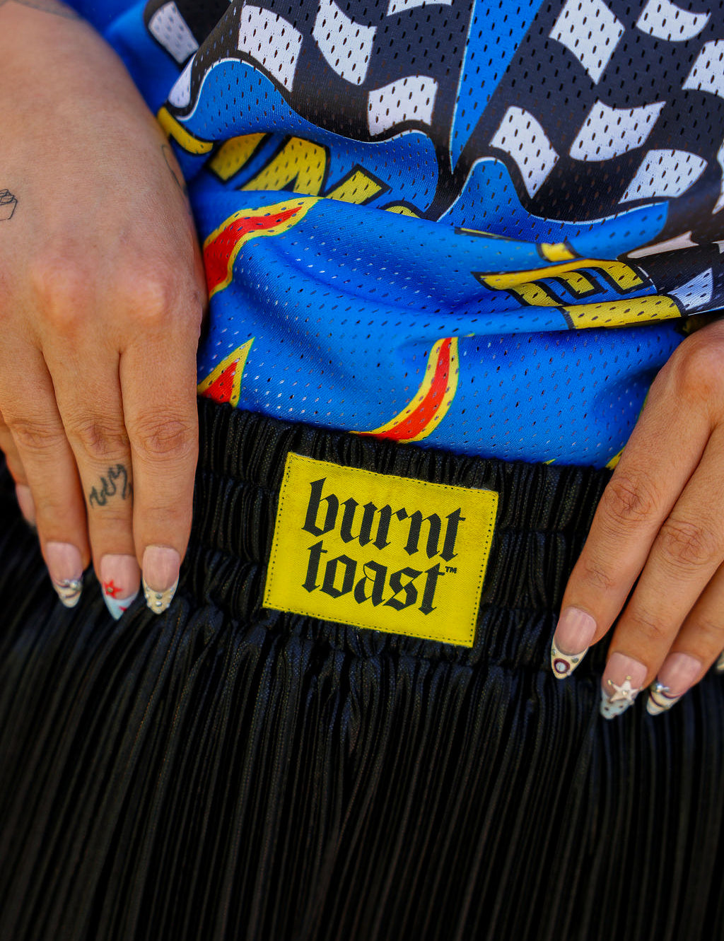 Close up of waistband of metallic black pleated shorts with a yellow burnt toast label blurred natural background, frontal view