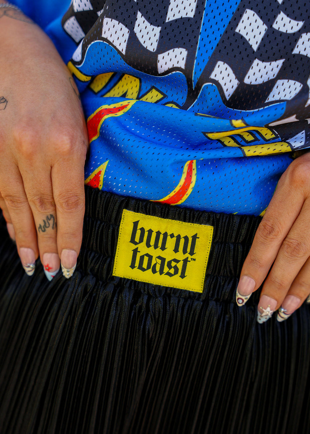 Close up of waistband of metallic black pleated shorts with a yellow burnt toast label blurred natural background, frontal view