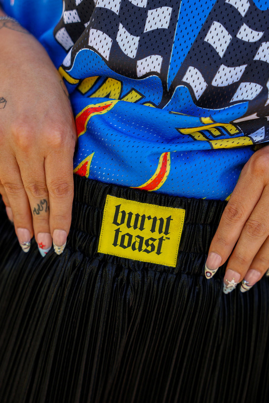Close up of waistband of metallic black pleated shorts with a yellow burnt toast label blurred natural background, frontal view
