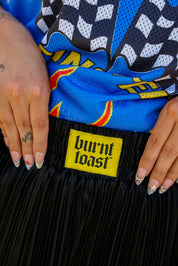 Close up of waistband of metallic black pleated shorts with a yellow burnt toast label blurred natural background, frontal view