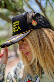 Model wearing a black trucker hat with 'God Bless America' gold text with red flame pin, outdoors, close up side view