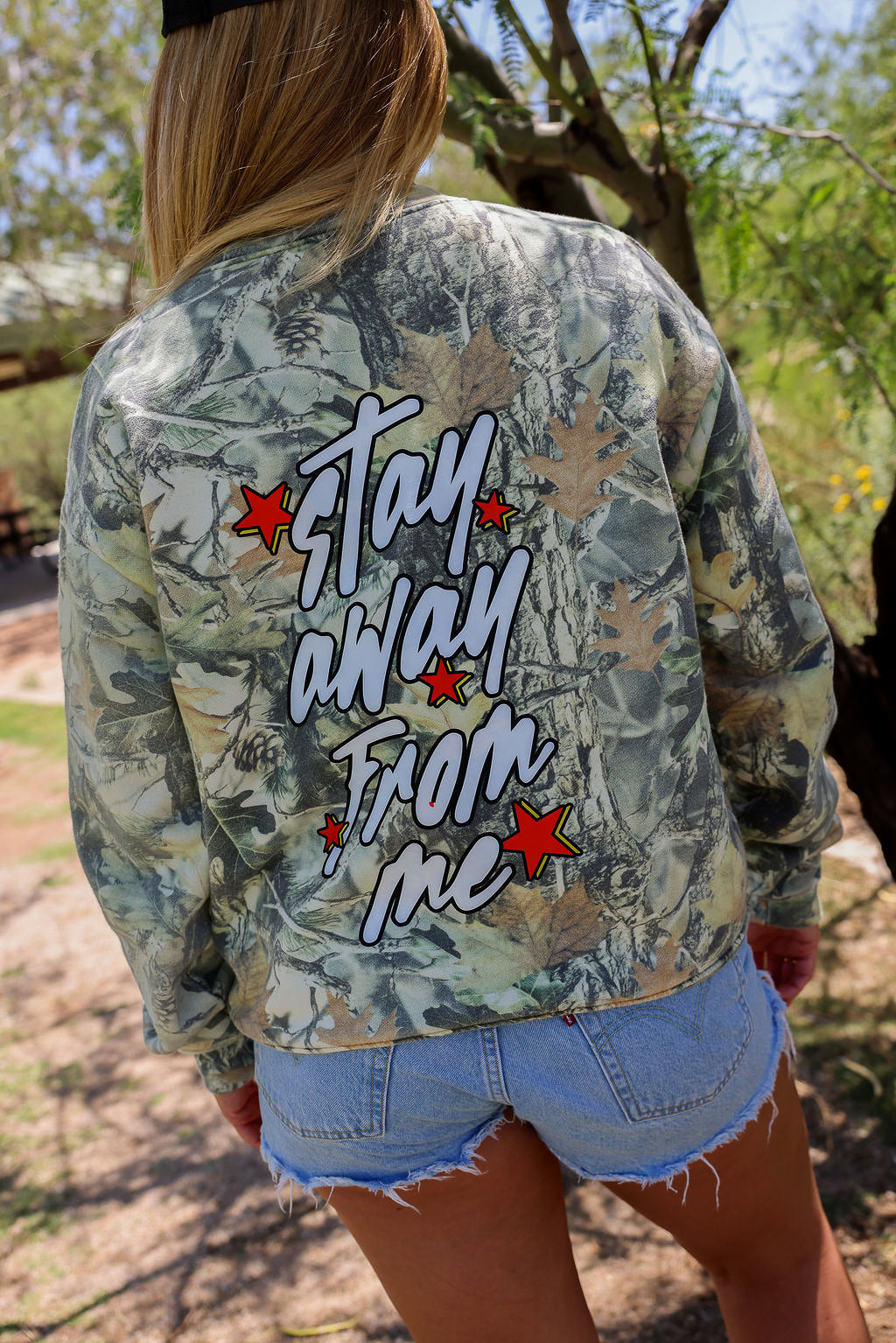 Person wearing a camouflage jacket with 'Stay Away From Me' text outdoors, back view