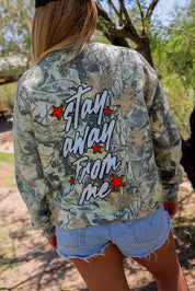 Person wearing a camouflage jacket with 'Stay Away From Me' text outdoors, back view