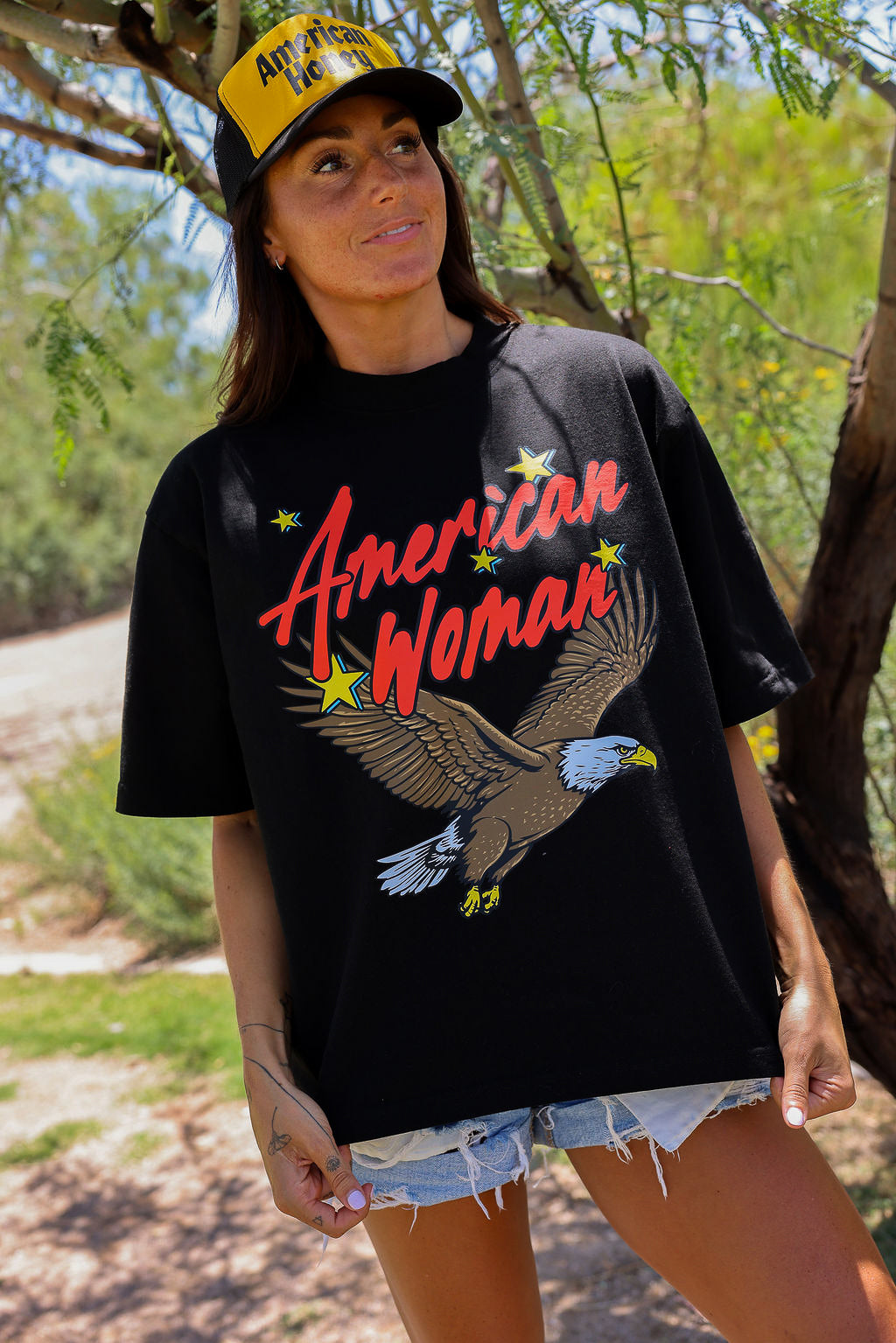 Woman wearing a black short sleeve shirt with 'American Woman' and an eagle graphic,  and yellow trucker hat, standing outdoors 
