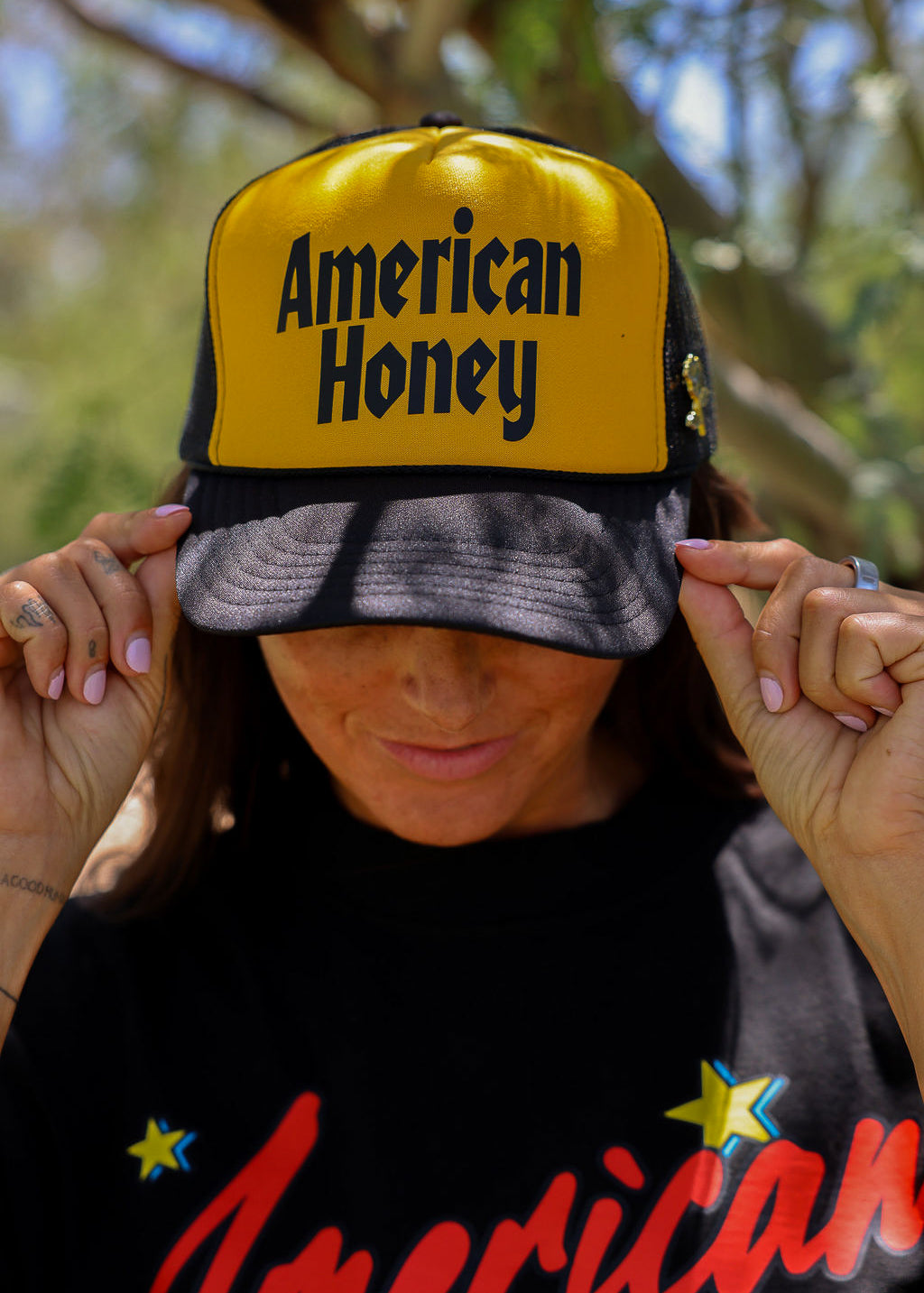 Person wearing a trucker at with 'American Honey' text and a black shirt with 'American' text, outdoors, close up frontal view