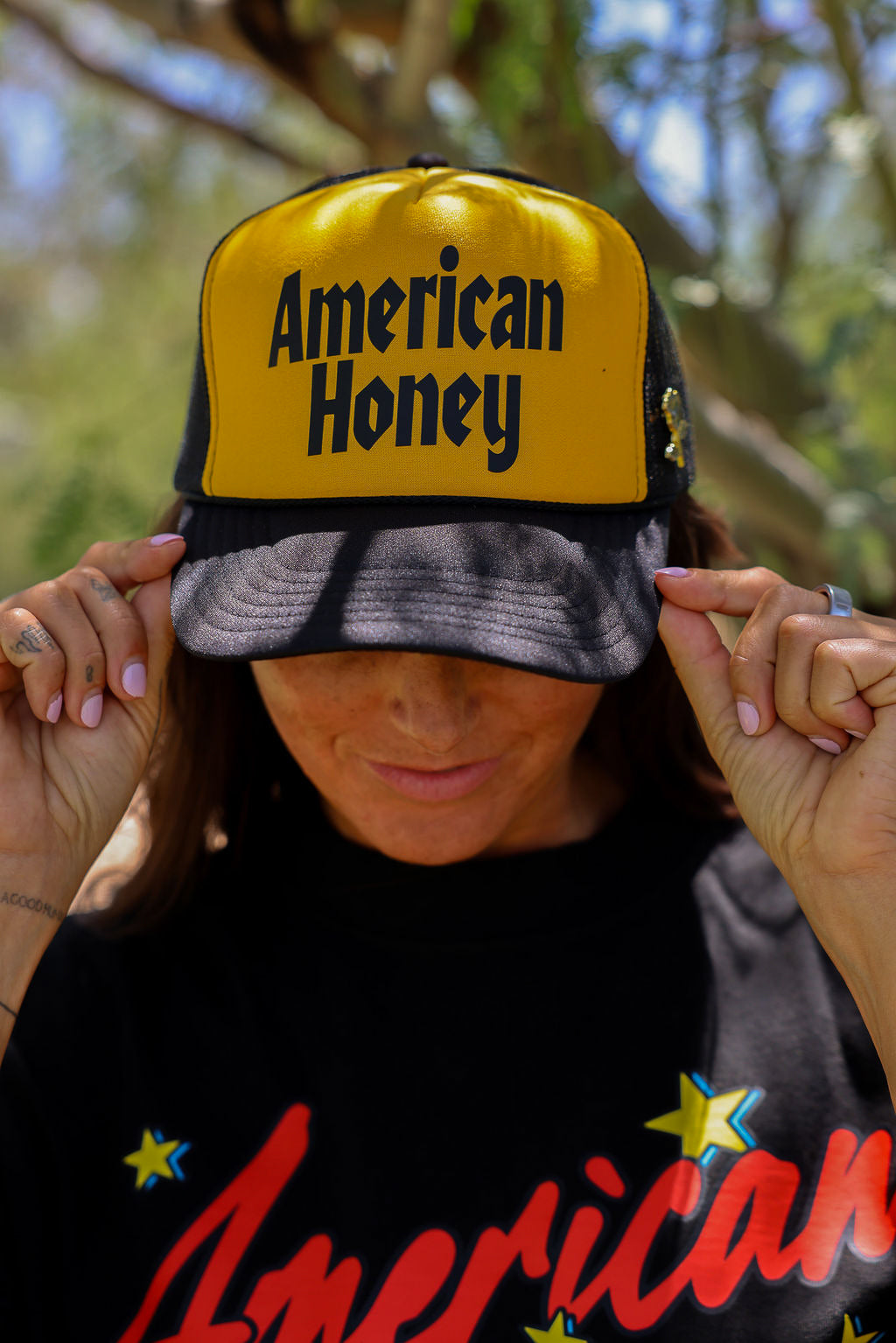 Person wearing a trucker at with 'American Honey' text and a black shirt with 'American' text, outdoors, close up frontal view