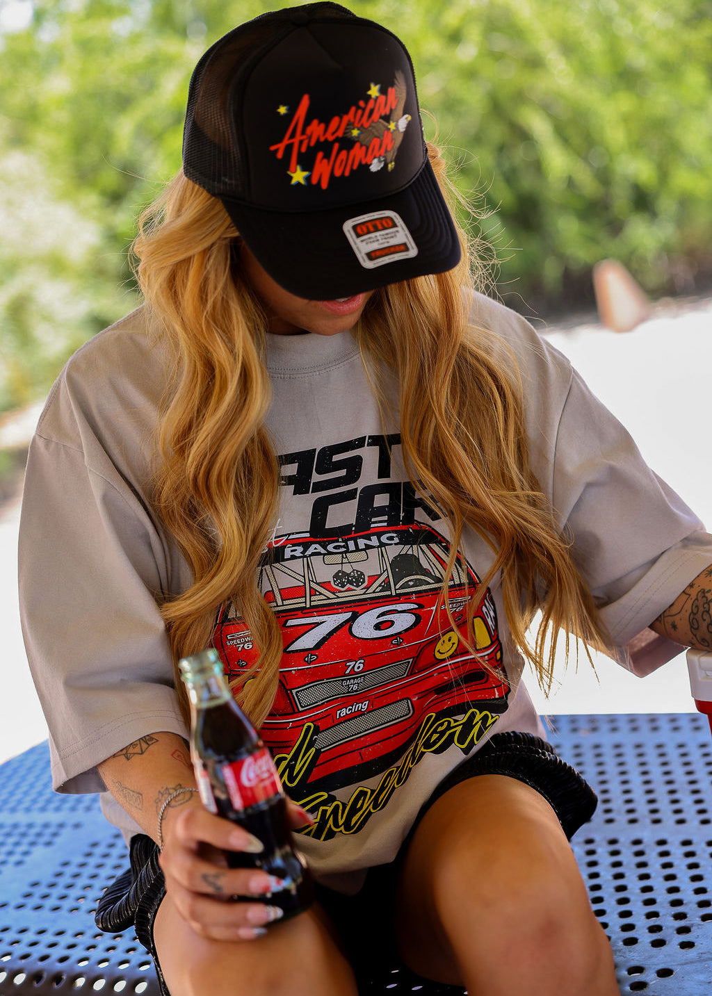 Model wearing a black trucker hat with American Woman text in red and t-shirt with racing design, holding a bottle outdoors, frontal view